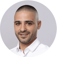 Man with short-cropped hair and a trimmed beard, identified as Nehorai Kadosh, wearing a white shirt, smiling slightly, and positioned against a plain light gray background.