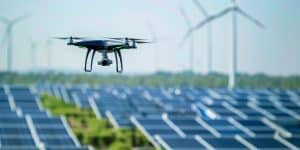 Aerial drone hovers over a solar panel field with wind turbines in the background.